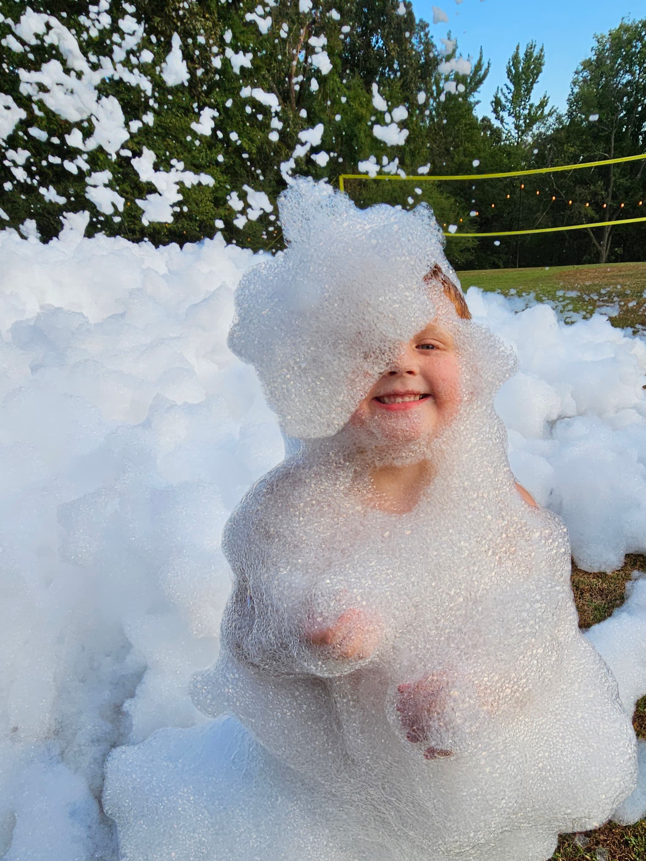 A smiling child is playfully covered in bubbles in a field.