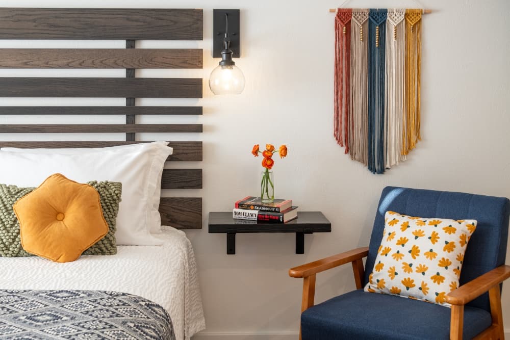 A cozy bedroom with a wooden headboard, adorned with decorative pillows, a side table with books and flowers, and a colorful wall hanging.