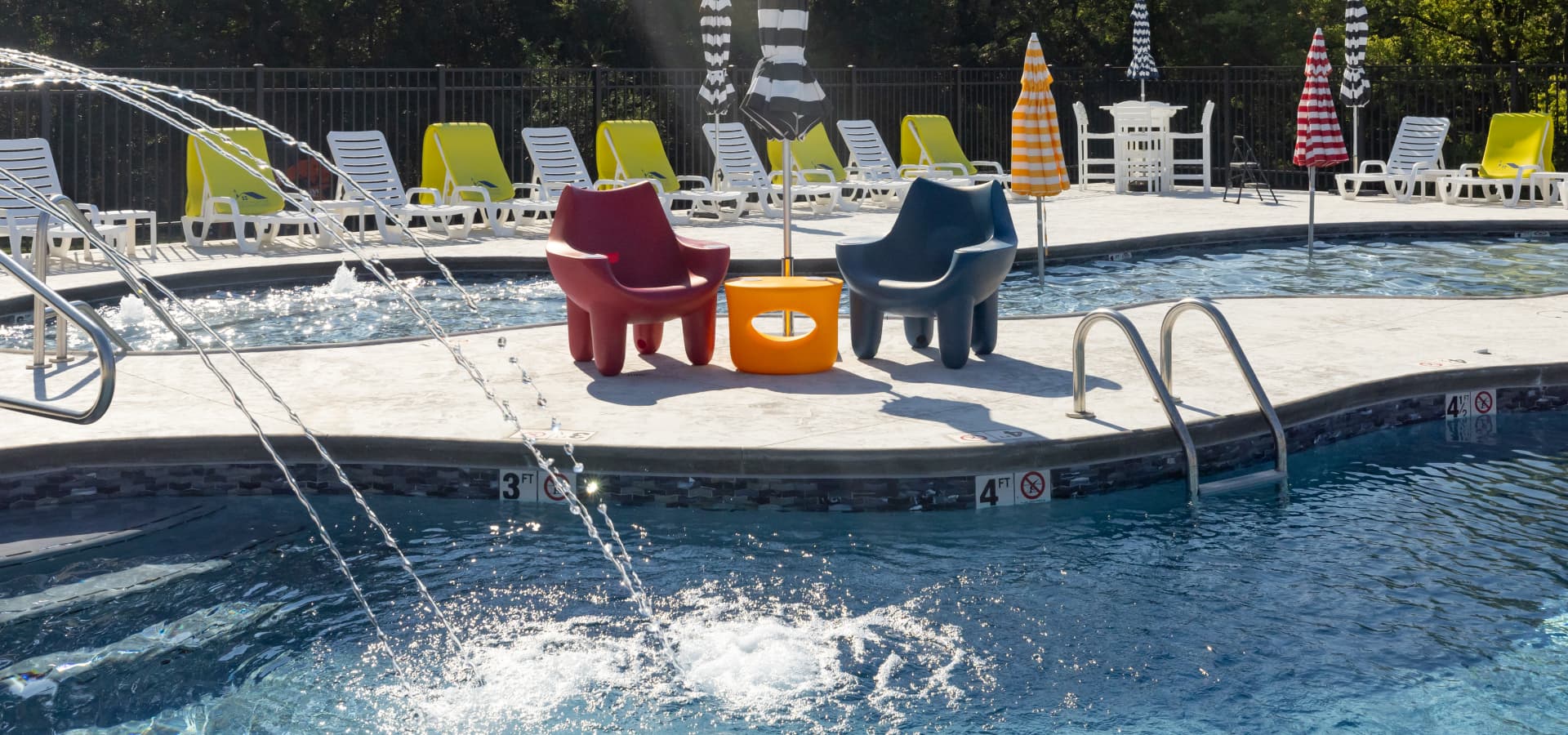 Two colorful chairs and a small table are on the edge of a swimming pool, surrounded by lounge chairs and umbrellas.