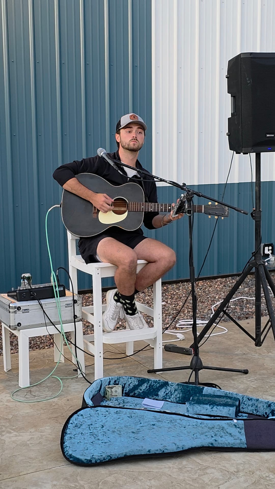 A musician performs on a stool with an acoustic guitar in an outdoor setting.