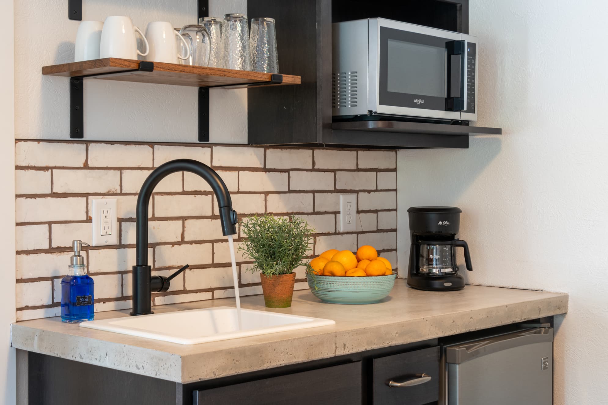 A modern kitchen countertop features a faucet, a bowl of oranges, a coffee maker, and a microwave above, set against a brick backsplash.